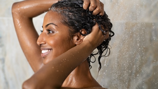 Woman washing hair showering in bathroom at home. Smiling black woman bathing while looking away. Happy woman rinsing hair while taking a shower at luxury spa.