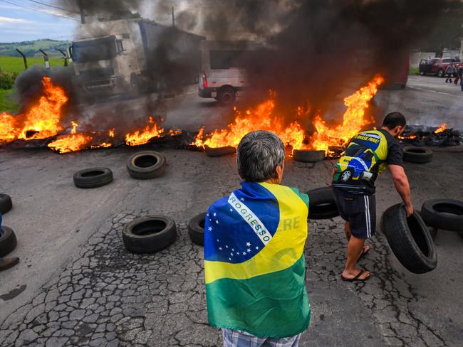 VARGINHA, BRAZIL - OCTOBER 31: A demonstrator wearing a Brazilian flag looks at a person putting tyres to make a burning barricade as truck drivers and supporters of President Jair Bolsonaro block 491 road to protest against the results of the presidential run-off on October 31, 2022 in Varginha, Brazil. Blockades have been registered in at least 11 states. Leftist leader Lula da Silva defeated incumbent Bolsonaro and will rule the country from January 2023. (Photo by Pedro Vilela/Getty Images)