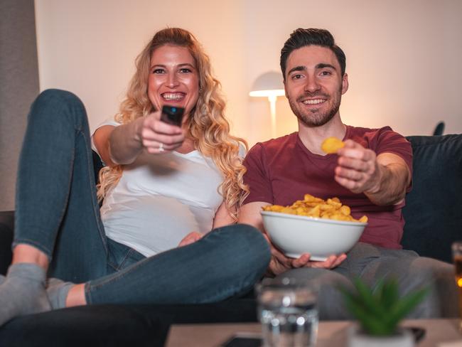 Happy young caucasian pregnant woman watching TV with her husband in the living room. They are eating snacks and drinking water and beer.