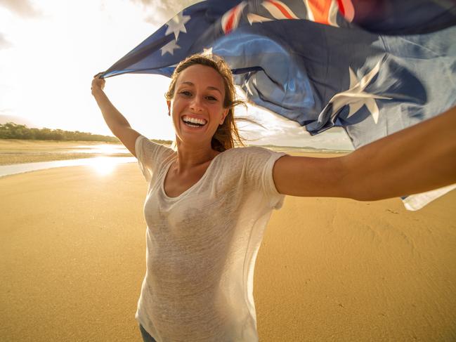 Young woman on the beach at sunset takes a selfie portrait while holding an Australian flag in the air.