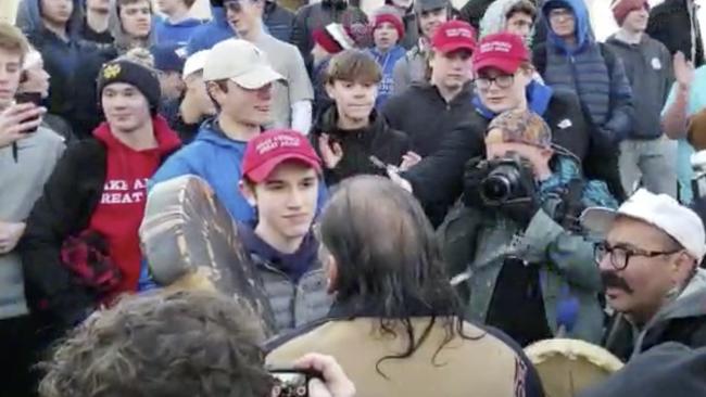 A teenager wearing a "Make America Great Again" hat stands sneering at an elderly Native American war vet singing and playing a drum in Washington. (Survival Media Agency via AP)