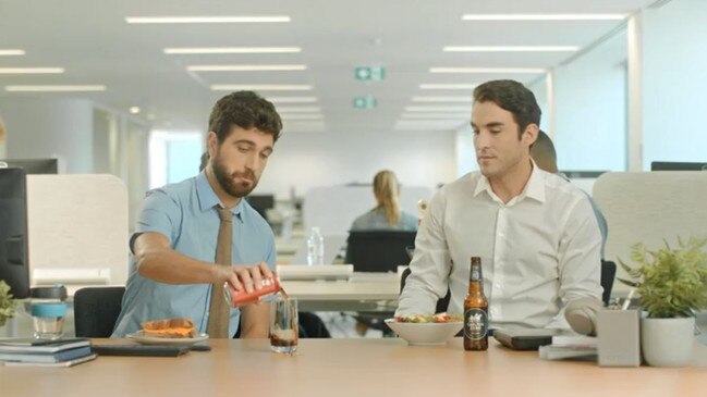 The ad features two male office workers at lunch. One has a bottle of non-alcoholic beer with a salad, while the other pours a can of sugary cola.