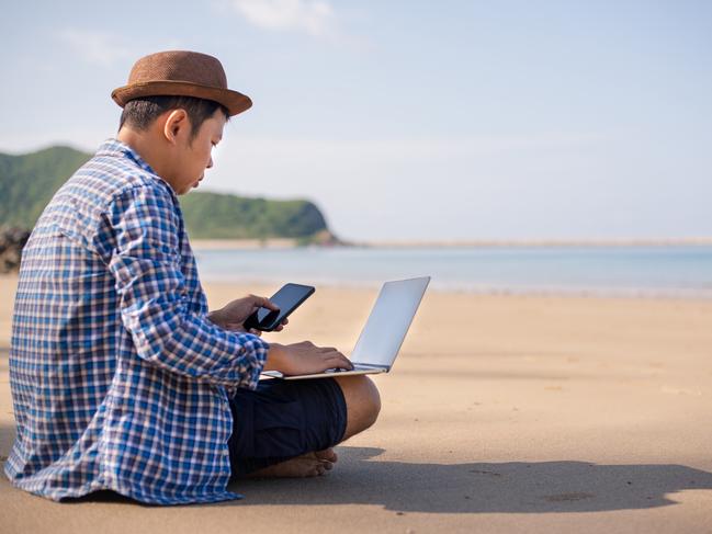 QLD JOBS - freelancer young man in a shirt and shorts and sneakers working on laptop with smartphone while sitting on the summer beach, sending mail,relax and vacation concept. Picture: Istock