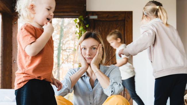 Frustrating woman alone struggling burnout with kids. Stressed out mother sitting on floor in middle of toys while children naughty running around her at room. Family home with chaos, mess.