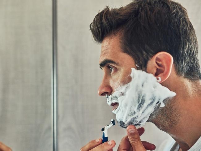 Shot of a focused young man shaving his beard while looking at his reflection in the mirror at home