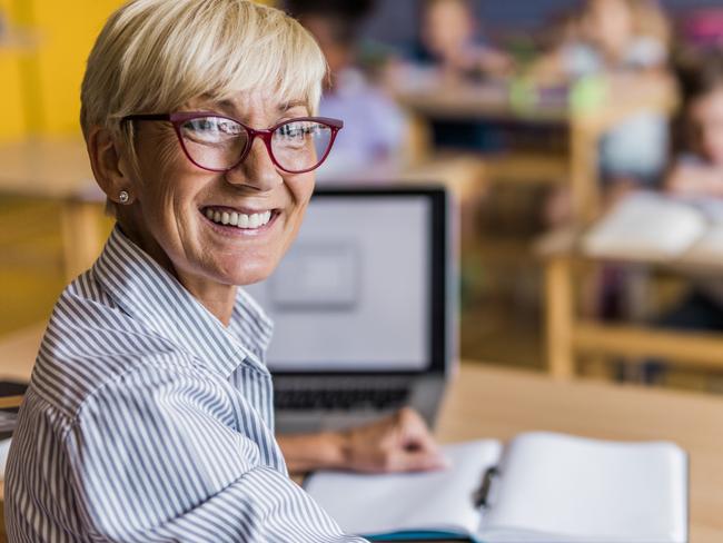 CAREERS: Happy senior teacher sitting on a class and looking at camera. Elementary students are in the background.