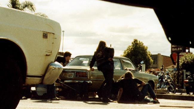 Comanchero bikie gang member Robert Heeney standing beside the car outside the line of fire at the Milperra Massacre, while injured John Hennessey is sitting, Glen Eaves crouches on the left with a shotgun, and Comanchero leader Jock Ross lies injured beneath the car.