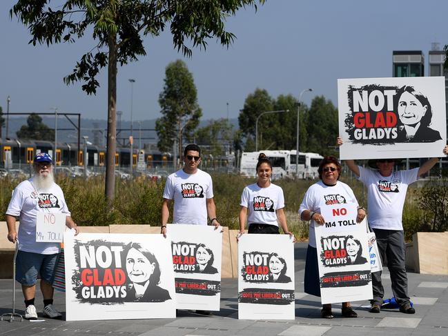 Protesters hold placards as Gladys Berejiklian and Andrew Constance speak to media during a press conference at Penrith Station. Picture: AAP