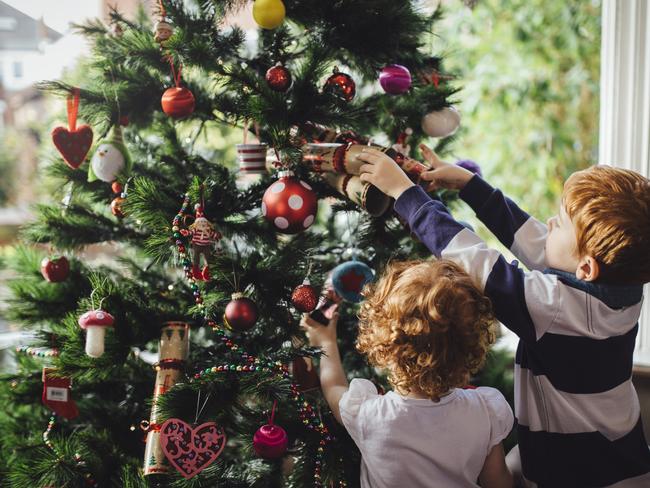 A young red head boy and his sister are decorating the Christmas tree in their home. He is reaching up and putting a bauble onto the tree while his sister tries to help.