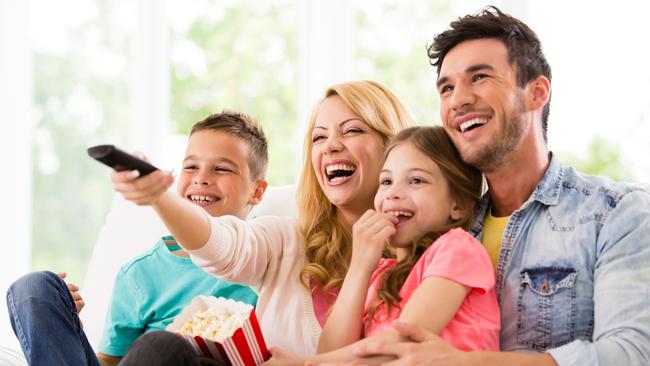 A happy family watches TV at home.  A dad in a denim shirt holds a little girl in a pink t-shirt.  A smiling mom holds a container of popcorn and directs a remote control toward a television.  A young boy wears a turquoise t-shirt and a pair of blue jeans.