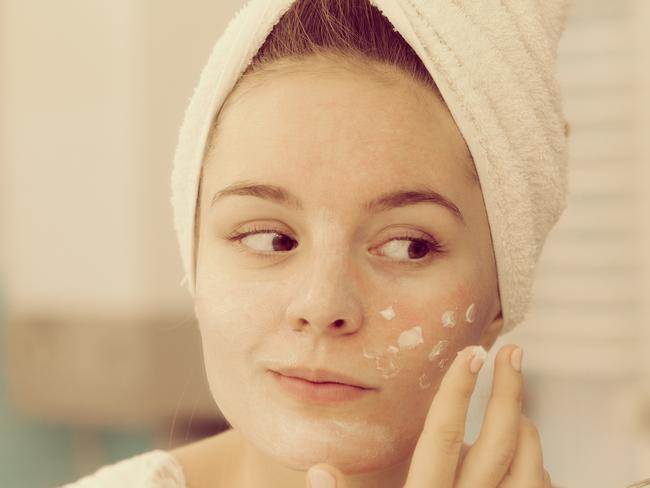 Woman applying mask moisturizing skin cream on face looking in bathroom mirror. Girl taking care of her complexion layering moisturizer. Skincare spa treatment.