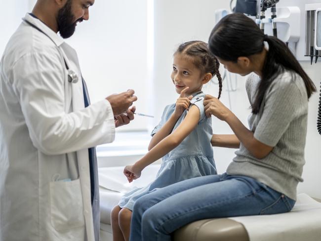 A young girl around 7 years old is at a doctor's office with her mother. They are both sitting on an examination table. She is looking at her doctor and pointing at her arm. The doctor is holding a syringe and ready to administer the flu shot onto his young patient's arm.