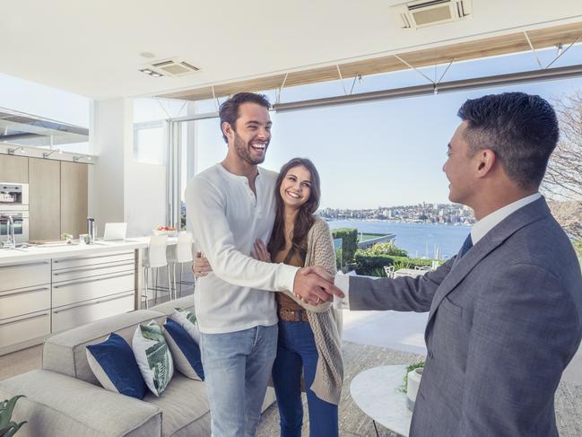 Real estate agent with couple in luxury home. They are shaking hands. There is a water view, kitchen and living room in the background. Couple are casually dressed. They are laughing. Agent is dressed in a suit and smiling. Wide angle. Picture: Istock