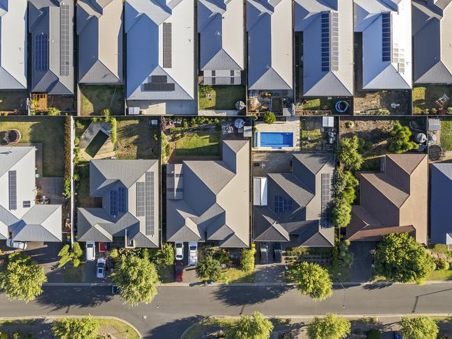 Aerial view directly above new outer suburban/semi-rural housing development with single-level housing between two streets with t-intersections and orange coloured street trees.  Mostly gray metal roofing, landscaped front and back yards, some solar panels, cars and motor vehicles parked in driveways and street, some street and parkland trees, water tanks in backyards, sheds and garages, swimming pools.  Mount Barker, South Australia; property investment housing money generic