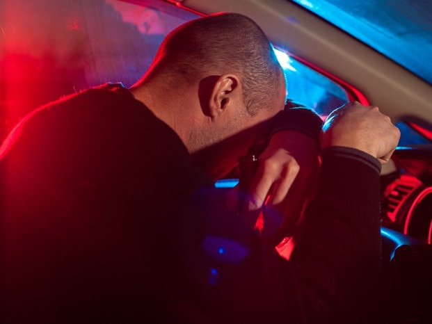 Upset drunk driver is caught driving under alcohol influence. Man covering his face from police car light. Photo: iStock