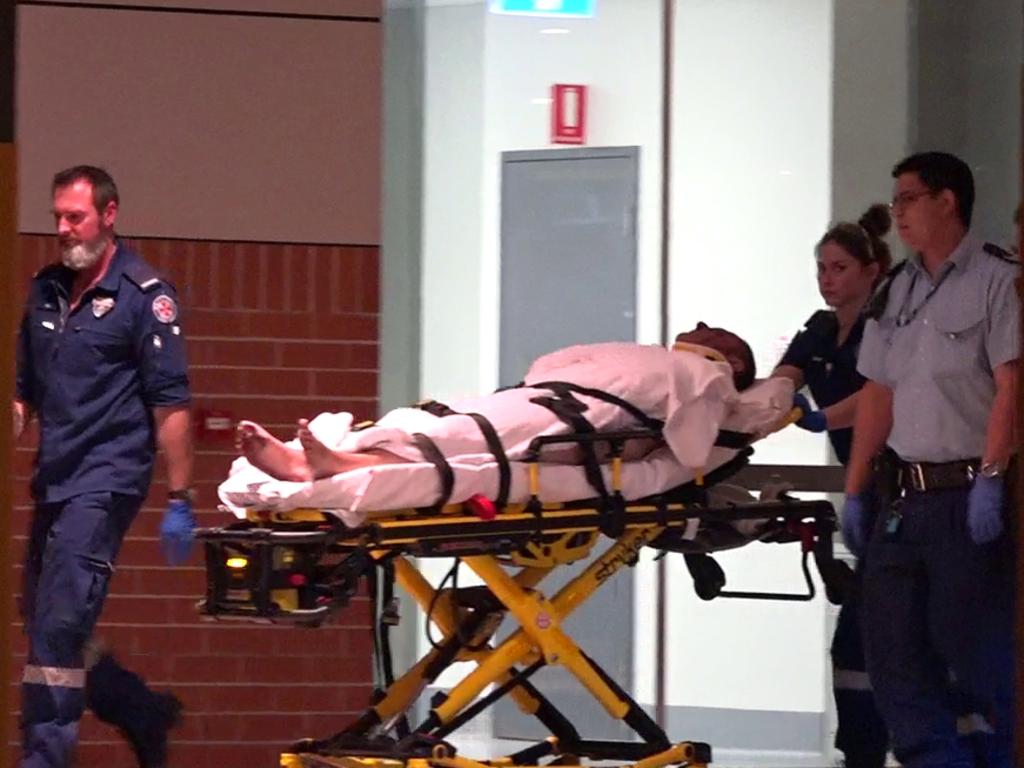 A man strapped on a gurney is taken to hospital after the alleged attack eleven days ago.