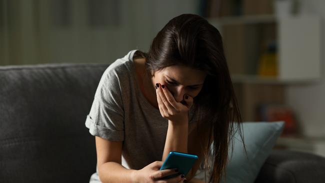 Sad teen receiving bad news online in a smart phone sitting on a couch in the living room at home