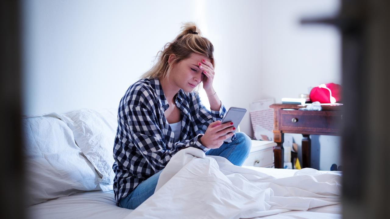 Young sad emotional pretty girl seated on bed with smartphone