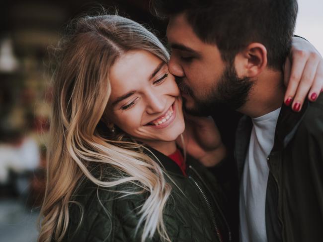 Precious moments of love. Close up portrait of handsome bearded guy kissing his girlfriend in cheek while she hugging him. Lady closing eyes with pleasure and smiling